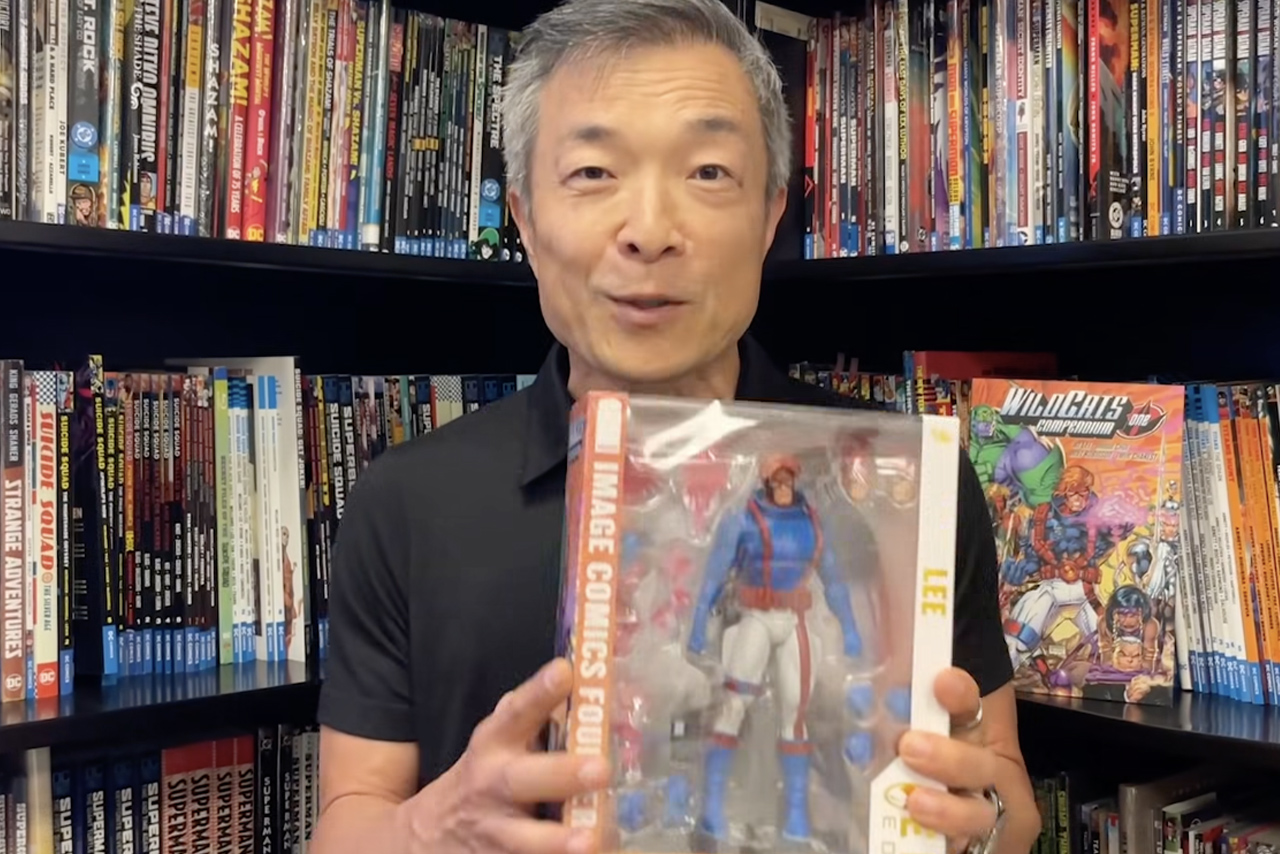 Jim Lee holding the packaged McFarlane Spartan WildC.A.T.s Elite Edition action figure in front of a comic book shelf.