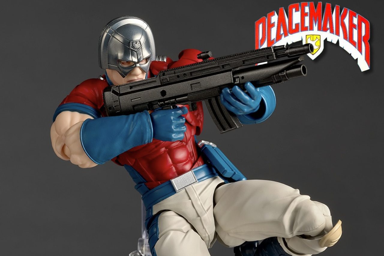 A close-up promotional photograph of the Kaiyodo Amazing Yamaguchi Peacemaker action figure. The figure, a man in a silver helmet, red and blue suit, and beige pants, is in a dynamic pose. He has a determined expression and is aiming a black assault rifle toward the right. The background is a solid grey. In the top right corner, a red-lettered logo with a white outline and eagle motif reads "PEACEMAKER".